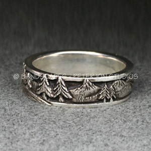 May include: A silver band ring featuring a relief design of a mountain range and pine trees around the exterior. The text BANDSCAPES - PETER JON STUDIOS is engraved along the center of the band. The metal has a polished finish on the edges.
