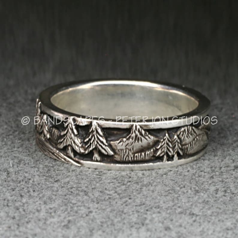 May include: A silver band ring featuring a relief design of a mountain range and pine trees around the exterior. The text BANDSCAPES - PETER JON STUDIOS is engraved along the center of the band. The metal has a polished finish on the edges.