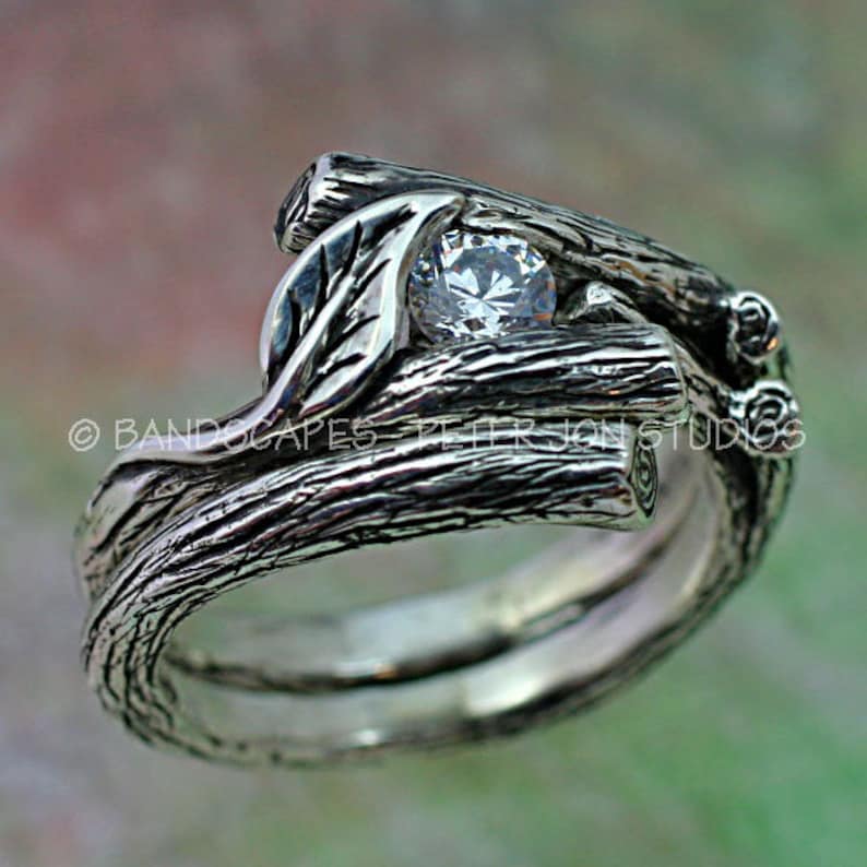 May include: A silver ring set with a single round diamond, the band is crafted to resemble intertwined branches with a leaf detail.