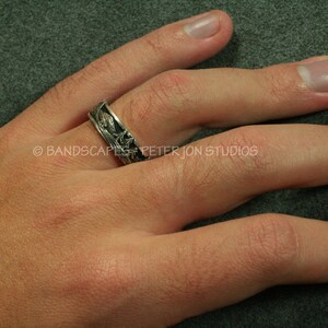 May include: A silver-toned metal ring featuring a detailed relief pattern of a landscape with trees and mountains. The band has a dark, oxidized finish in the recessed areas to highlight the engraved scenery. Text reads BANDSCAPES PETER JON STUDIOS.