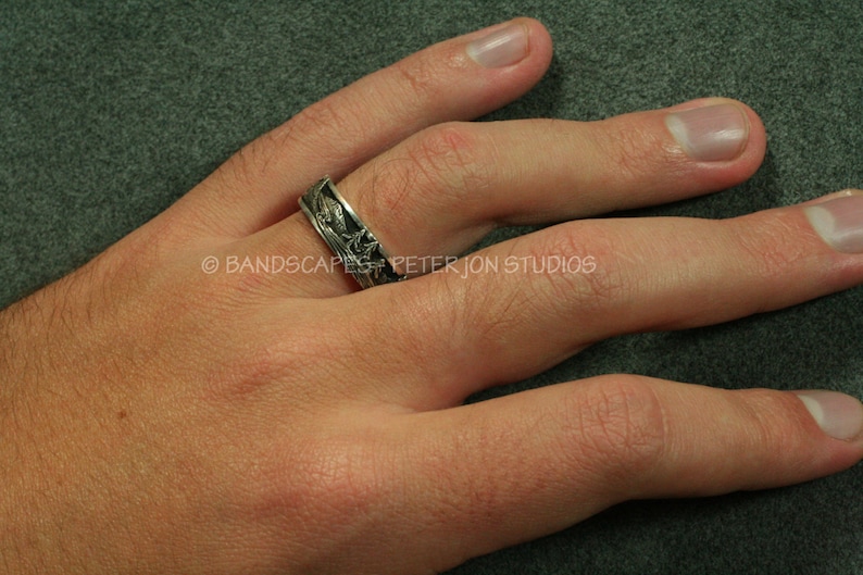 May include: A silver-toned metal ring featuring a detailed relief pattern of a landscape with trees and mountains. The band has a dark, oxidized finish in the recessed areas to highlight the engraved scenery. Text reads BANDSCAPES PETER JON STUDIOS.