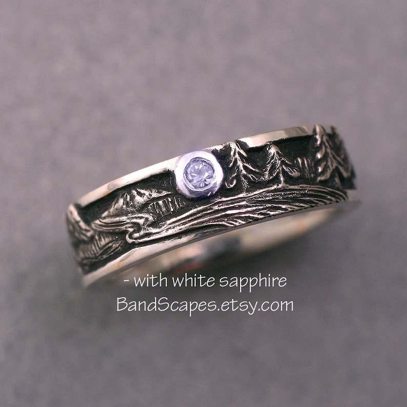 May include: A silver ring featuring a textured landscape relief of trees and mountains with a single bezel-set white sapphire. The ring includes the text - with white sapphire BandScapes.etsy.com printed on the lower portion of the image.