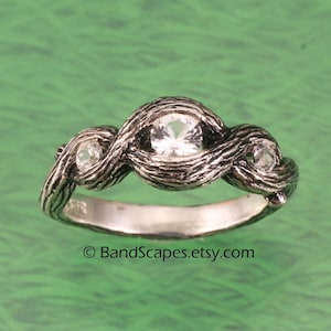 Könnte beinhalten: Ein silberfarbener Ring mit drei klaren, runden Edelsteinen. Das Ringband hat ein strukturiertes, organisches Design, das verschlungenen Ästen ähnelt. Der Etsy-Shop-Name "BandScapes" ist sichtbar.