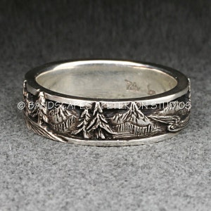 May include: A silver band ring featuring a detailed relief carving of a mountain landscape with evergreen trees. The exterior surface displays a continuous scene of peaks and forest. The text BANDSCAPES A PETERION STUDIOS is visible across the center.