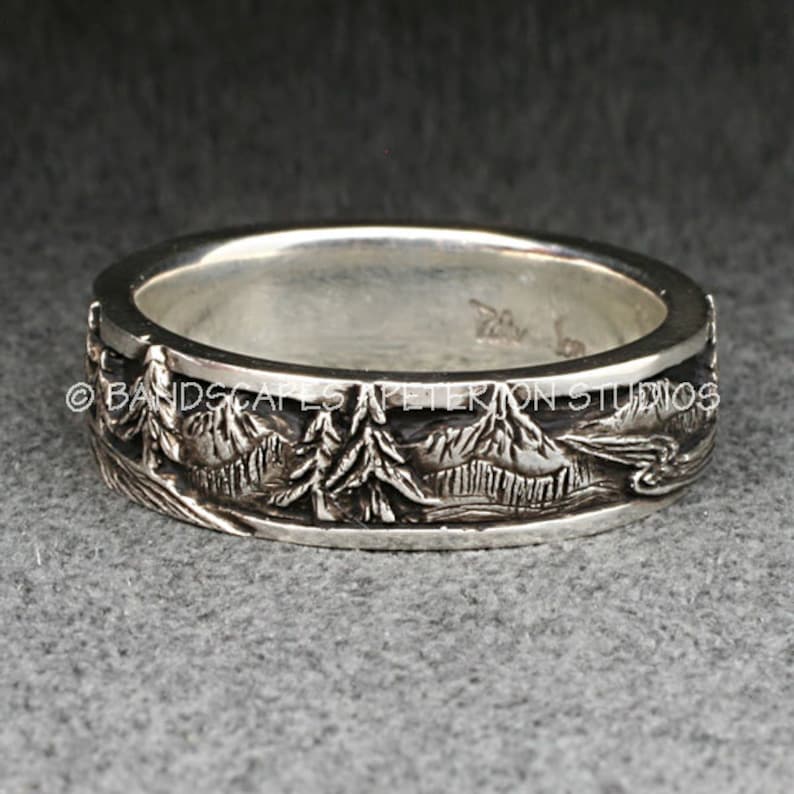 May include: A silver band ring featuring a detailed relief carving of a mountain landscape with evergreen trees. The exterior surface displays a continuous scene of peaks and forest. The text BANDSCAPES A PETERION STUDIOS is visible across the center.