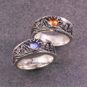 May include: Two silver rings with a mountain range design. The ring on the left has a blue gemstone, and the ring on the right has an orange gemstone. © BandScapes.etsy.com