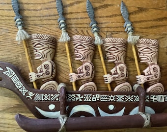 May include: A carved wooden outrigger canoe with four tiki figures holding spears. The canoe is brown with white and cream-colored decorative patterns. The tiki figures are beige with carved details.