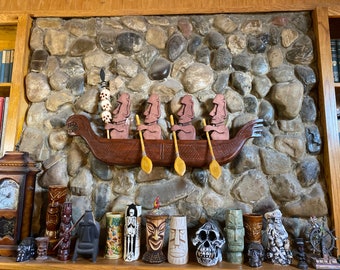 May include: A wooden wall carving depicts four Moai statues in a canoe, with paddles. Above, a stone wall serves as a backdrop. Below, a collection of tiki mugs and decorative items are displayed on a shelf, showcasing a Polynesian theme.