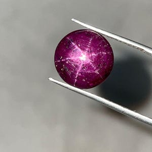 Könnte beinhalten: Ein runder, polierter Rubin-Edelstein, der von einer Pinzette gehalten wird. Der Edelstein hat eine tiefe, satte magenta Farbe mit einem sechsstrahligen Sterneffekt. Der Stern ist hellweiß und kontrastiert mit dem dunklen Hintergrund des Steins.