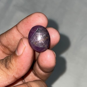 May include: A polished, oval-shaped cabochon gemstone with a deep purple hue. The stone displays a distinct six-rayed star pattern, known as asterism, visible on its smooth, rounded surface. The gem has a translucent quality with internal inclusions.