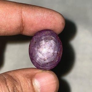 Peut inclure: Une pierre précieuse polie, de forme ovale, rouge-violet. La gemme présente un motif en forme d'étoile à sa surface. La pierre est tenue entre deux doigts, mettant en valeur sa taille et sa couleur. La gemme est un rubis.