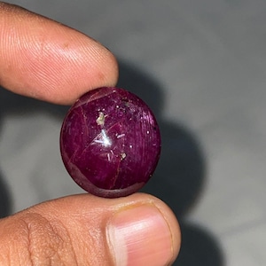 Peut inclure: Une pierre précieuse rubis taillée en cabochon rond, d'une couleur rouge magenta profond. La surface de la pierre présente un phénomène optique en forme d'étoile, appelé astérisme, avec une lumière réfléchie sur la surface polie.