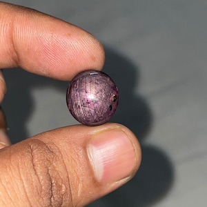 Puede incluir: Una piedra preciosa de rubí estrella tallada en cabujón redondo con un tono rojo violáceo intenso. La piedra muestra un efecto de estrella de seis rayos en su superficie pulida y abovedada, con inclusiones naturales y fibras visibles.