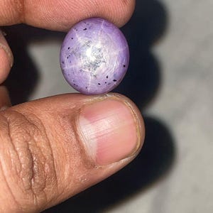 Könnte beinhalten: Ein polierter, oval geformter, violetter Edelstein mit sternförmigem Muster. Der Edelstein hat eine glatte Oberfläche mit kleinen schwarzen Einschlüssen und einem silbernen Zentrum. Der Edelstein wird zwischen zwei Fingern gehalten.