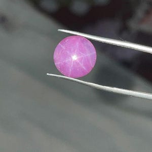Könnte beinhalten: Ein runder, rosa Edelstein mit sternförmigem Effekt, gehalten von einer silbernen Pinzette. Der Edelstein hat eine glatte Oberfläche und ein sechszackiges Sternmuster. Der Hintergrund ist unscharf, wodurch der Fokus auf dem Edelstein liegt.