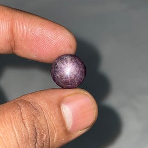 Puede incluir: Una piedra preciosa de rubí estrella de forma redonda, tallada en cabujón, con un tono rojo violáceo intenso. La piedra muestra un efecto de estrella de seis rayos, conocido como asterismo, visible en su superficie pulida y abovedada.