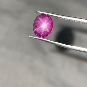 Pode incluir: Uma gema de rubi oval, segurada por pinças prateadas. O rubi, de cor rosa intensa, exibe um efeito de estrela de seis pontas. A gema é lisa e polida, refletindo a luz. O fundo é cinza neutro.