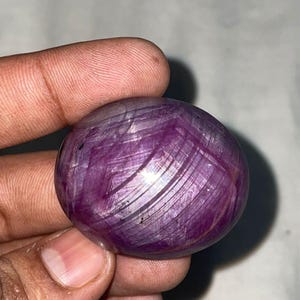May include: A polished, round gemstone with a deep purple hue. The stone has a swirling, fibrous pattern throughout, with lighter streaks creating visual depth. The gem is held between two fingers, showcasing its size and texture.