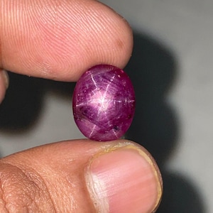 Puede incluir: Gema ovalada de color rojo intenso con un efecto de estrella de seis rayos. La superficie pulida refleja la luz, creando un patrón de estrella. La gema se sostiene entre dos dedos, mostrando su color y forma.