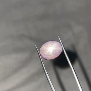 May include: A light purple, oval gemstone held by silver tweezers. The gem has a pearlescent sheen and a subtle, textured surface. The background is a solid, muted grey.