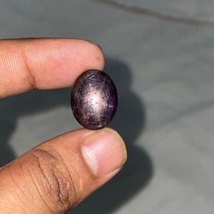 Könnte beinhalten: Ein ovaler Cabochon-Edelstein mit einem tiefvioletten und dunkelroten Farbton. Der Stein weist ein ausgeprägtes sechsstrahliges Sternmuster auf seiner polierten, gewölbten Oberfläche auf, das durch Lichtreflexion an internen Einschlüssen entsteht.