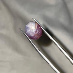 May include: A round, polished gemstone held by silver tweezers. The gem is a mix of purple and white, with a star-like effect visible on its surface. The gem is likely a star ruby, a type of corundum.