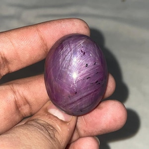 May include: A polished, oval-shaped gemstone in shades of deep purple and magenta. The stone has a star-like pattern across its surface, with dark inclusions. The gem is held in a hand, showcasing its size and color.