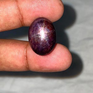 May include: An oval-shaped cabochon star ruby gemstone with a deep purple-red hue. The stone features a distinct six-rayed star effect, known as asterism, visible on its polished, smooth surface, reflecting light in a centered, intersecting pattern.