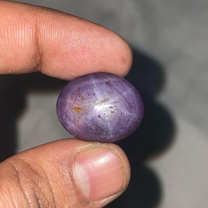 Pode incluir: Uma pedra preciosa redonda e polida, de cor roxa, com um padrão em forma de estrela. A gema é segurada entre dois dedos, mostrando sua forma esférica e variações de cor. A pedra tem uma superfície lisa com inclusões naturais.