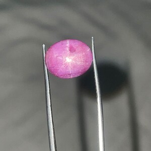 May include: A pink, oval gemstone held by tweezers. The gem has a slightly translucent appearance with internal light reflections. The tweezers are silver, and the background is a blurred grey.