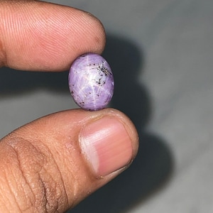 May include: An oval-shaped, cabochon-cut star sapphire gemstone in a light purple hue. The stone features a visible six-rayed star effect across its surface, with dark, natural inclusions scattered throughout the translucent, polished material.