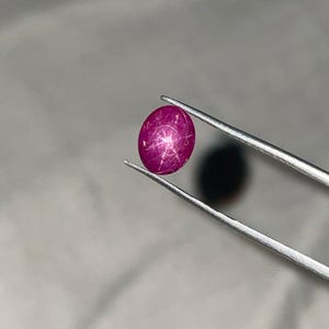 Puede incluir: Una gema de rubí ovalada, sujeta con pinzas. La gema exhibe un efecto de estrella, con líneas que irradian desde el centro. El rubí es de un color rojo intenso, y está sobre un fondo neutro. Las pinzas son plateadas.