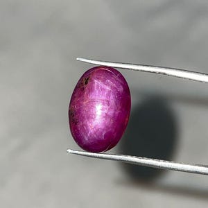 Könnte beinhalten: Ein polierter, ovalförmiger Rubin-Edelstein, der von einer Pinzette gehalten wird. Der Edelstein hat eine tiefe, lebendige, rötlich-rosa Farbe mit einer glatten, reflektierenden Oberfläche. Der Edelstein ist etwa 1,9 cm lang.