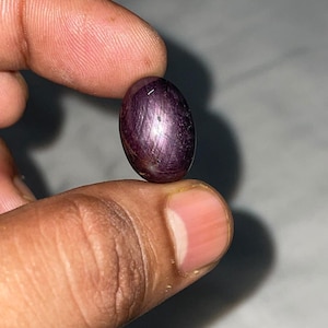 Può includere: Una gemma di rubino stellato di forma ovale, color viola intenso, con una superficie cabochon liscia e lucida. La pietra mostra un distinto effetto a stella a sei raggi sul suo corpo scuro e traslucido, riflettendo la luce.