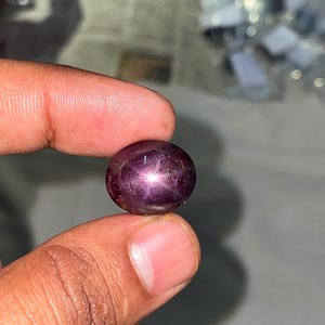 Puede incluir: Una piedra preciosa de rubí estrella de color púrpura intenso y forma ovalada, con un distintivo efecto de estrella de seis rayos en su superficie pulida en cabujón. La gema muestra un color rico y saturado con inclusiones naturales visibles.