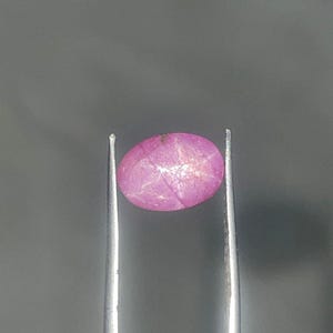 Peut inclure: Une pierre précieuse ovale rose, tenue par une pince argentée, sur fond gris. La pierre présente un motif en forme d'étoile à sa surface, avec des nuances de rose et de blanc. La pince est positionnée verticalement.