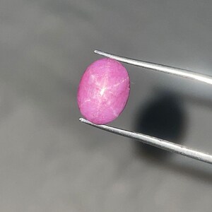 Puede incluir: Una gema rosa pulida, de forma ovalada, con un patrón en forma de estrella. La gema está sujeta por unas pinzas plateadas sobre un fondo gris. El efecto de estrella es visible en la superficie de la piedra.