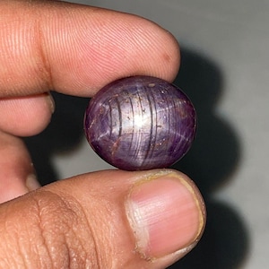 以下が含まれることがあります： 星の効果がある、丸く磨かれた紫色の宝石。宝石は指の間に挟まれ、滑らかで反射する表面を持っています。星の効果は、石の中のインクルージョンに光が反射することによって生まれます。
