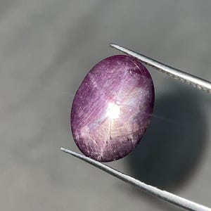 Könnte beinhalten: Ein polierter, oval geformter Edelstein, der von einer Pinzette gehalten wird. Der Edelstein hat eine tiefviolette Farbe mit einem sternartigen Effekt auf seiner Oberfläche. Der Sterneneffekt wird durch Lichtreflexionen von Einschlüssen im Stein verursacht. Der Hintergrund ist ein neutrales Grau.