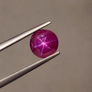 Op de afbeelding: Een ronde, cabochon geslepen sterrobijn edelsteen met een diepe magenta-rode kleur. De steen vertoont een duidelijk zesstralig asterisme-effect, dat verschijnt als een helder wit sterpatroon in het midden van het gepolijste, gladde oppervlak.