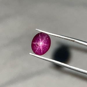 Może przedstawiać: Żywy, owalny kamień szlachetny rubinu, trzymany przez pęsetę. Klejnot ma głęboki, purpurowy kolor z sześcioramiennym wzorem gwiazdy widocznym na jego powierzchni. Kamień szlachetny jest centralnym punktem, prezentując jego naturalne piękno i unikalny efekt gwiazdy.