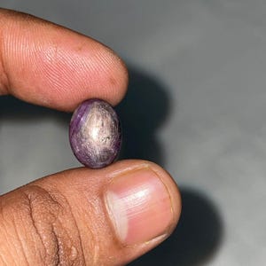 Può includere: Una gemma a cabochon di forma ovale con una profonda tonalità viola e magenta, caratterizzata da un distinto effetto a stella a sei raggi sulla superficie lucida. La pietra presenta una parte superiore liscia e arrotondata.