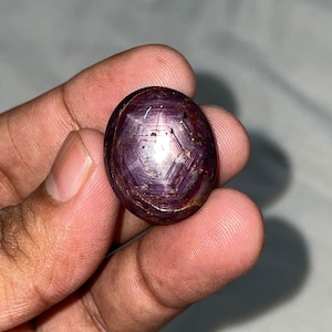 Pode incluir: Uma pedra preciosa em formato de cabochão oval com um tom roxo avermelhado profundo. A pedra exibe um padrão distinto de estrela de seis pontas em sua superfície polida e abobadada, criado pela luz refletida nas inclusões internas.