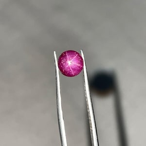 以下が含まれることがあります： ピンセットで保持された、丸く磨かれたスターサファイアの宝石。 ルビーは、表面に六条の星模様が見える、深いピンクがかった赤色をしています。 ピンセットは銀色で、背景はぼやけたグレーです。
