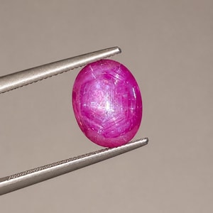 May include: An oval-shaped, cabochon-cut star ruby gemstone with a vibrant magenta-pink hue. The stone displays a distinct six-rayed asterism effect across its polished surface, held securely by a pair of metallic silver-toned tweezers.