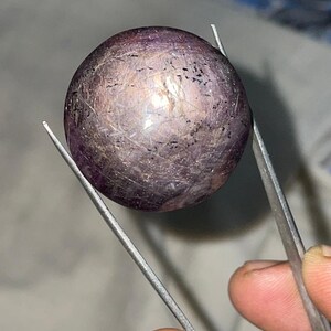 Può includere: Una gemma rotonda e lucidata tenuta da una pinzetta. La pietra è di un colore viola intenso con sottili variazioni e una superficie riflettente. La gemma sembra avere inclusioni naturali e un aspetto leggermente strutturato. La pinzetta è color argento.