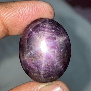 May include: A polished, oval-shaped gemstone with a deep purple hue. The stone exhibits a star effect, with lines radiating from a central point. The surface reflects light, creating a shimmering appearance. The gem is held between fingers.