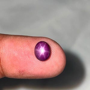 以下が含まれることがあります： 深いマゼンタ色をした小さな楕円形のスタールビーの宝石。カボションカットが施された滑らかで光沢のある表面には、アステリズムとして知られる鮮明な6条の星模様が中央の明るい光の反射として浮かび上がっています。