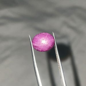 以下が含まれることがあります： 丸く磨かれたマゼンタ色の宝石が、銀色のピンセットで挟まれています。宝石はわずかにテクスチャ加工された表面を持ち、ぼやけた灰色の背景に配置されています。この宝石はルビーのようです。