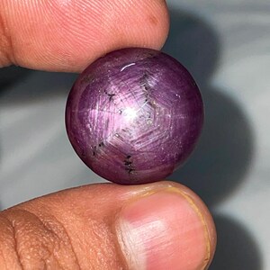 Op de afbeelding: Een ronde, in cabochon geslepen ster-robijn edelsteen met een diepe paarsrode kleur. De steen vertoont een duidelijk zesstralig asterisme-effect over het gepolijste oppervlak, met zichtbare natuurlijke insluitingen en een gladde vorm.